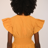 A person shown from the back wearing a mustard yellow peplum top with ruffled short sleeves and a smocked waist, paired with dark brown pants, against a plain off-white background.