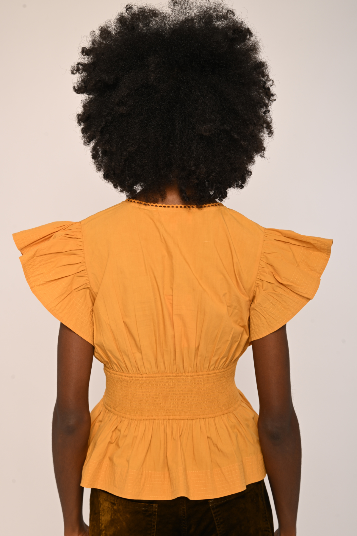 A person shown from the back wearing a mustard yellow peplum top with ruffled short sleeves and a smocked waist, paired with dark brown pants, against a plain off-white background.