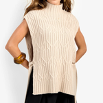 A woman modeling a sleeveless, high-neck cable-knit sweater vest in cream beige, paired with black pants, against a plain light gray studio background.