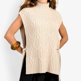 A woman modeling a sleeveless, high-neck cable-knit sweater vest in cream beige, paired with black pants, against a plain light gray studio background.
