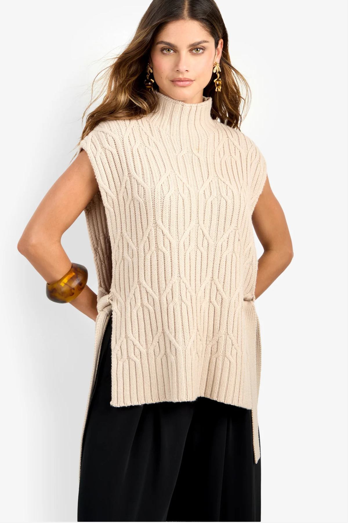 A woman modeling a sleeveless, high-neck cable-knit sweater vest in cream beige, paired with black pants, against a plain light gray studio background.