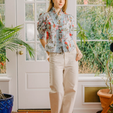 Woman in a floral blouse and beige pants standing in a room with plants and a door.