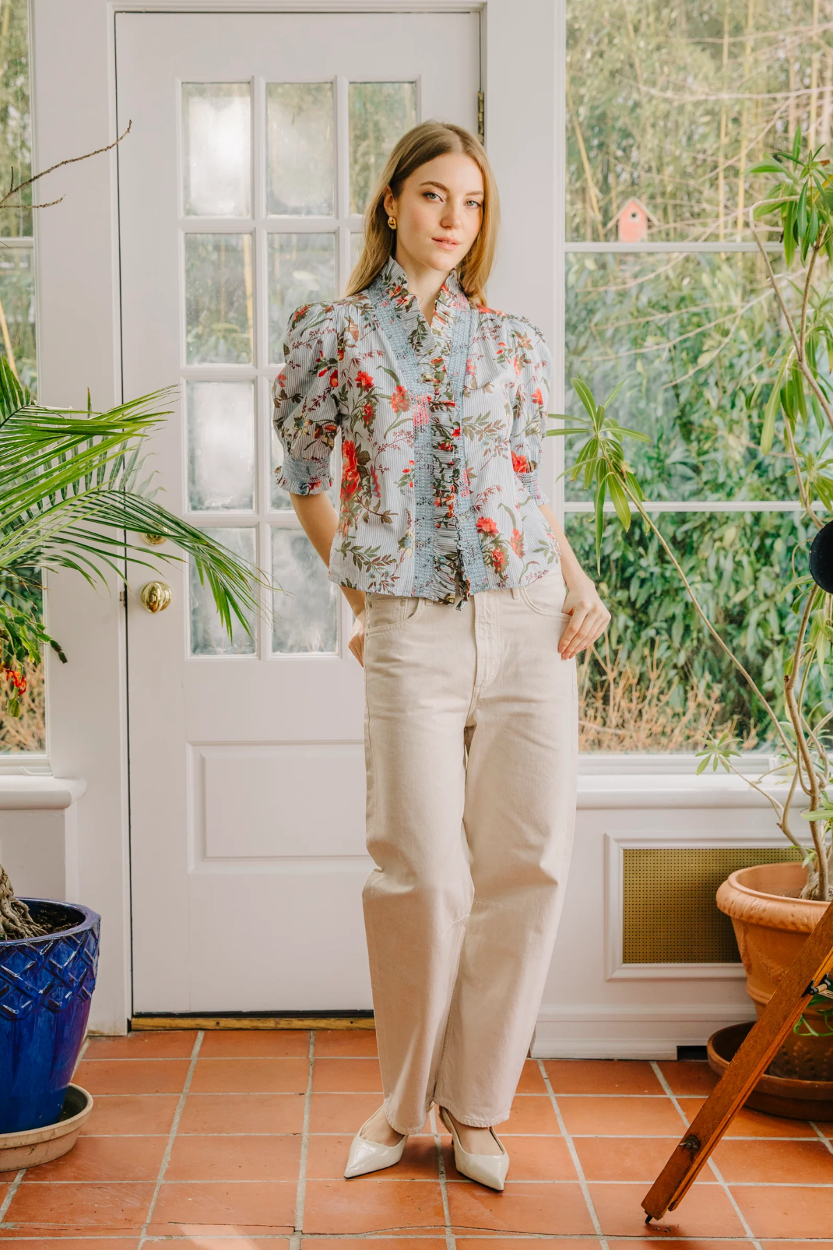 Woman in a floral blouse and beige pants standing in a room with plants and a door.