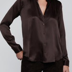 A woman wearing a dark brown satin button-up shirt and matching dark brown pants, shown from the mouth down against a light gray studio background.