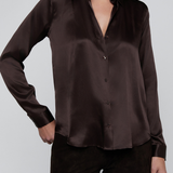 A woman wearing a dark brown satin button-up shirt and matching dark brown pants, shown from the mouth down against a light gray studio background.