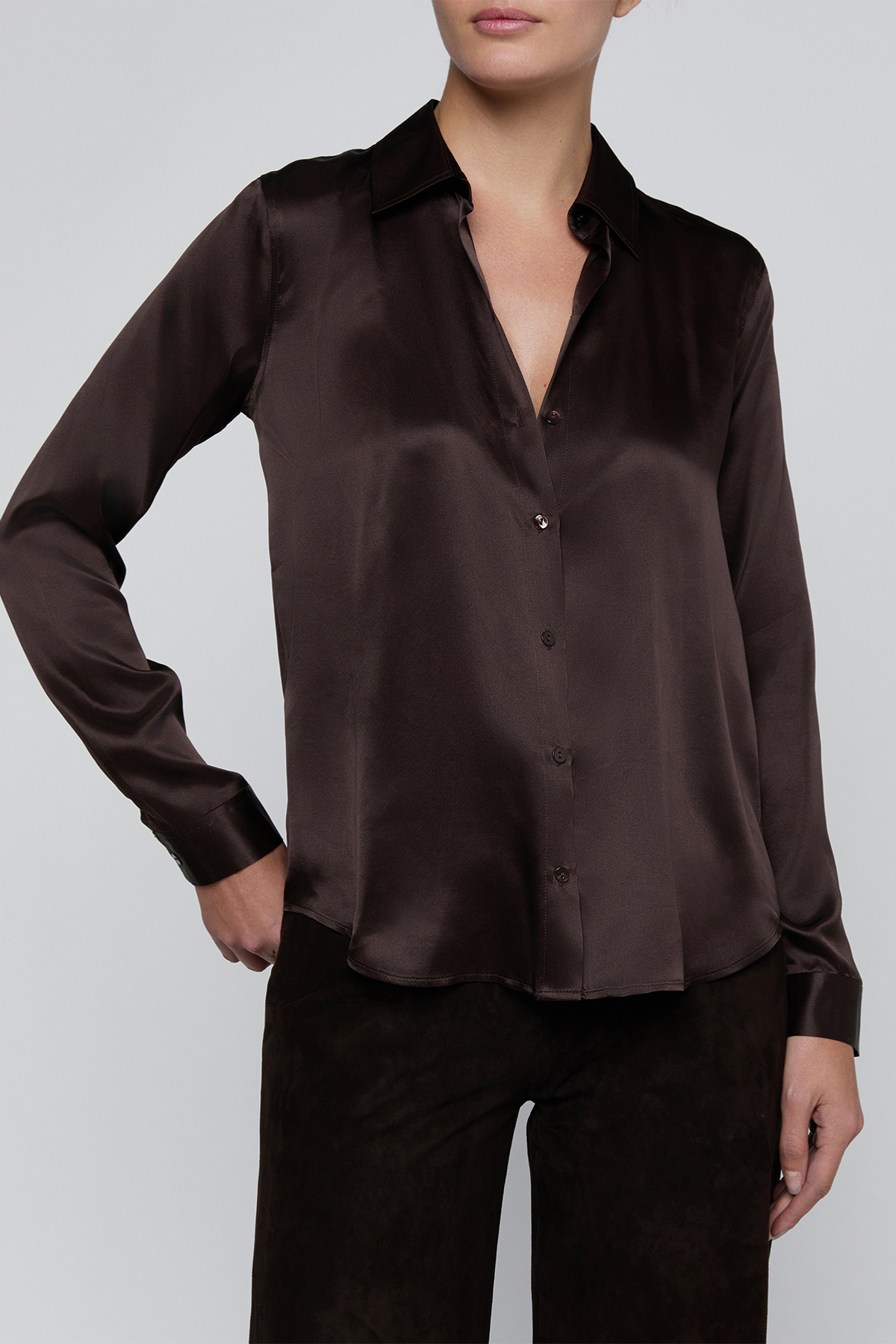 A woman wearing a dark brown satin button-up shirt and matching dark brown pants, shown from the mouth down against a light gray studio background.