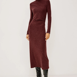 A model wearing a long-sleeve, high-neck ribbed knit midi dress in a deep burgundy color, paired with black knee-high leather boots, against a plain light gray studio background.
