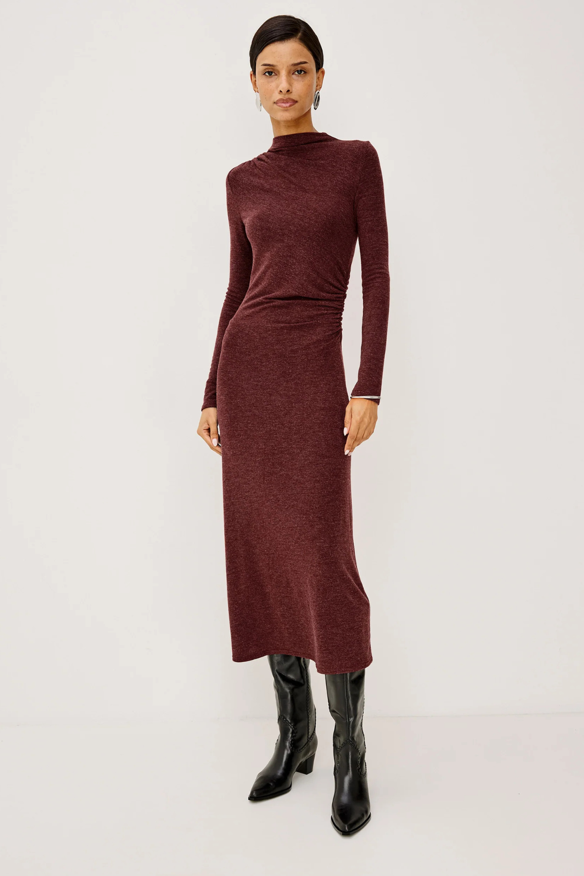 A model wearing a long-sleeve, high-neck ribbed knit midi dress in a deep burgundy color, paired with black knee-high leather boots, against a plain light gray studio background.