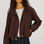 A woman modeling a dark brown cropped zip-up jacket with a drawstring hem, worn over a white t-shirt and dark blue jeans, against a light gray studio background.