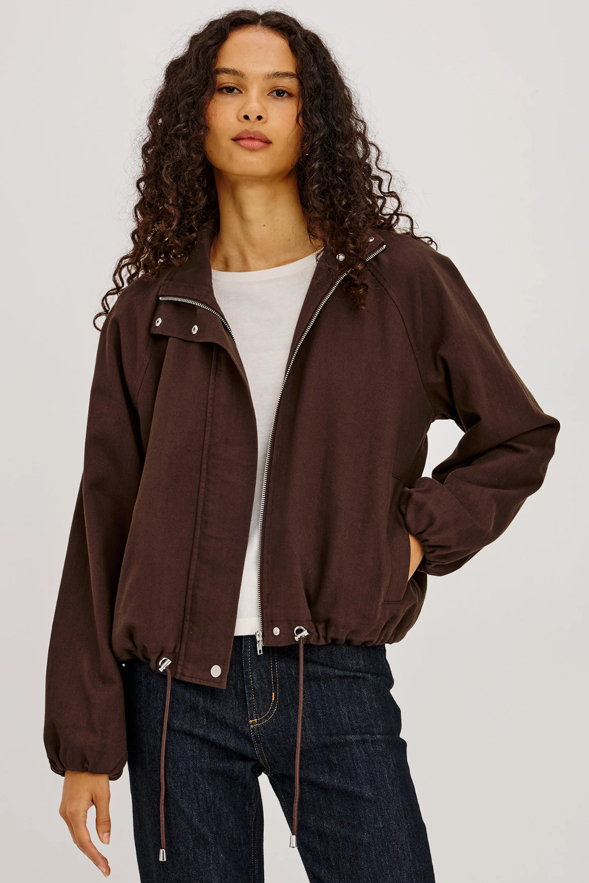 A woman modeling a dark brown cropped zip-up jacket with a drawstring hem, worn over a white t-shirt and dark blue jeans, against a light gray studio background.