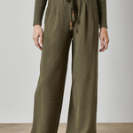 A person wearing olive green wide-leg ribbed knit pants with a drawstring waist, paired with a matching ribbed long-sleeve top and brown shoes, standing against a plain light gray wall and concrete floor.