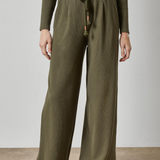 A person wearing olive green wide-leg ribbed knit pants with a drawstring waist, paired with a matching ribbed long-sleeve top and brown shoes, standing against a plain light gray wall and concrete floor.