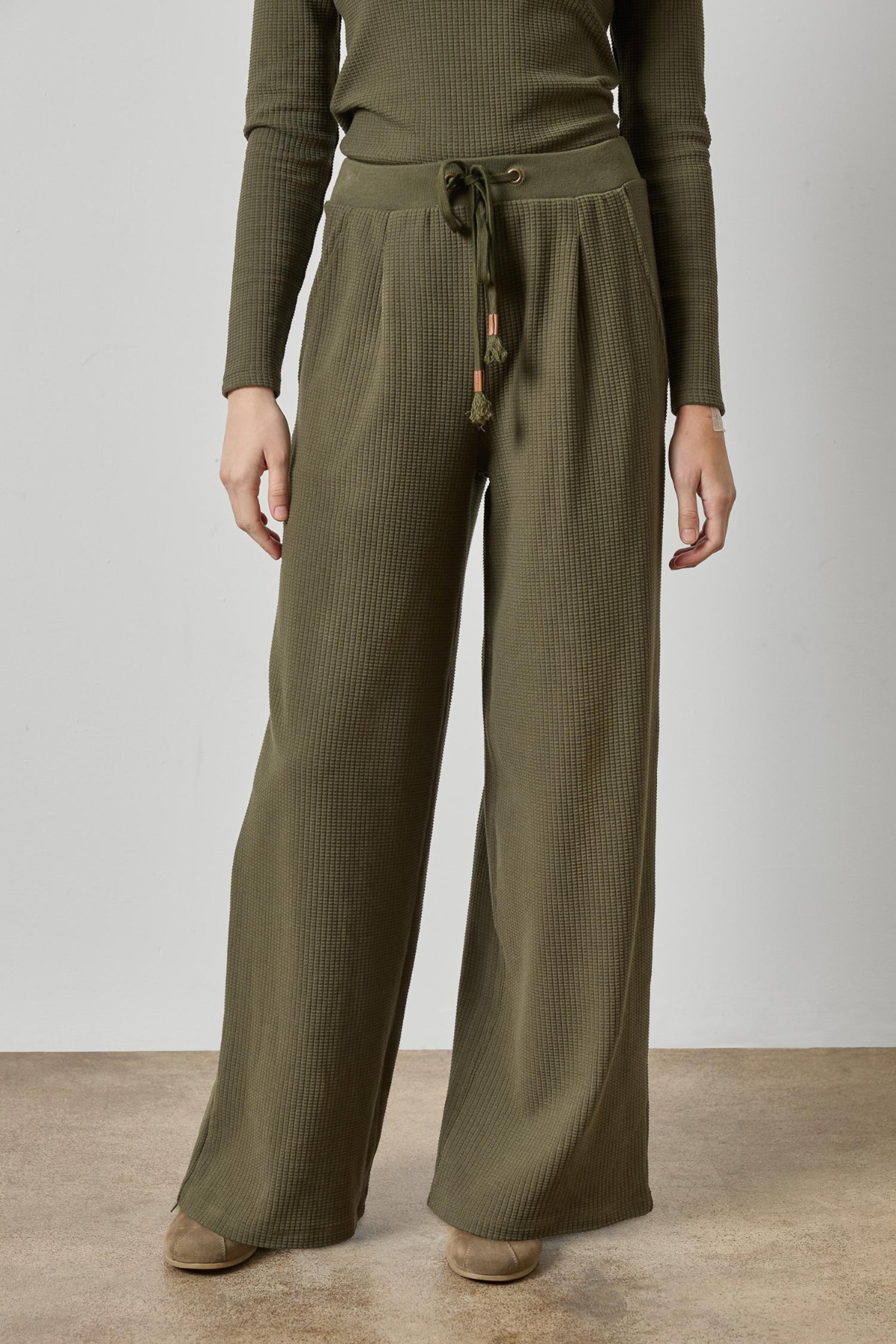 A person wearing olive green wide-leg ribbed knit pants with a drawstring waist, paired with a matching ribbed long-sleeve top and brown shoes, standing against a plain light gray wall and concrete floor.