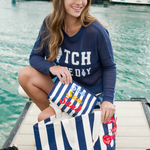 Woman sitting on a dock by water, holding a striped bag, wearing a blue cap and navy sweater with white text.