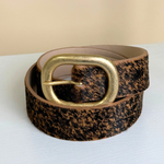 A textured leather belt with a brown and black speckled pattern and a large gold-tone buckle, coiled on a white surface against a beige background.