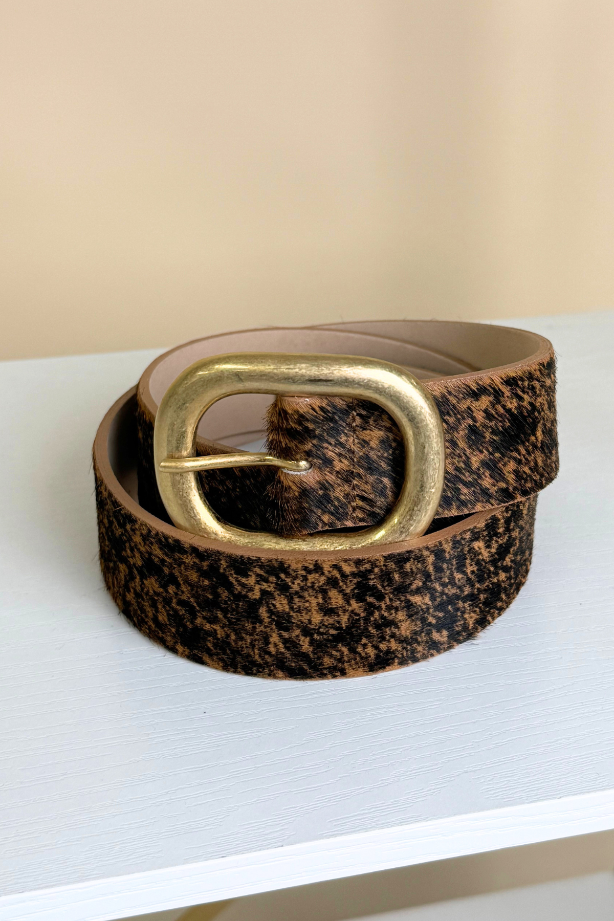 A textured leather belt with a brown and black speckled pattern and a large gold-tone buckle, coiled on a white surface against a beige background.