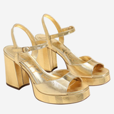 A pair of metallic gold platform block-heel sandals with open toes and ankle straps, shown on a light gray background.