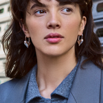 Woman wearing a gray blazer and blue shirt with earrings, standing in an urban setting.