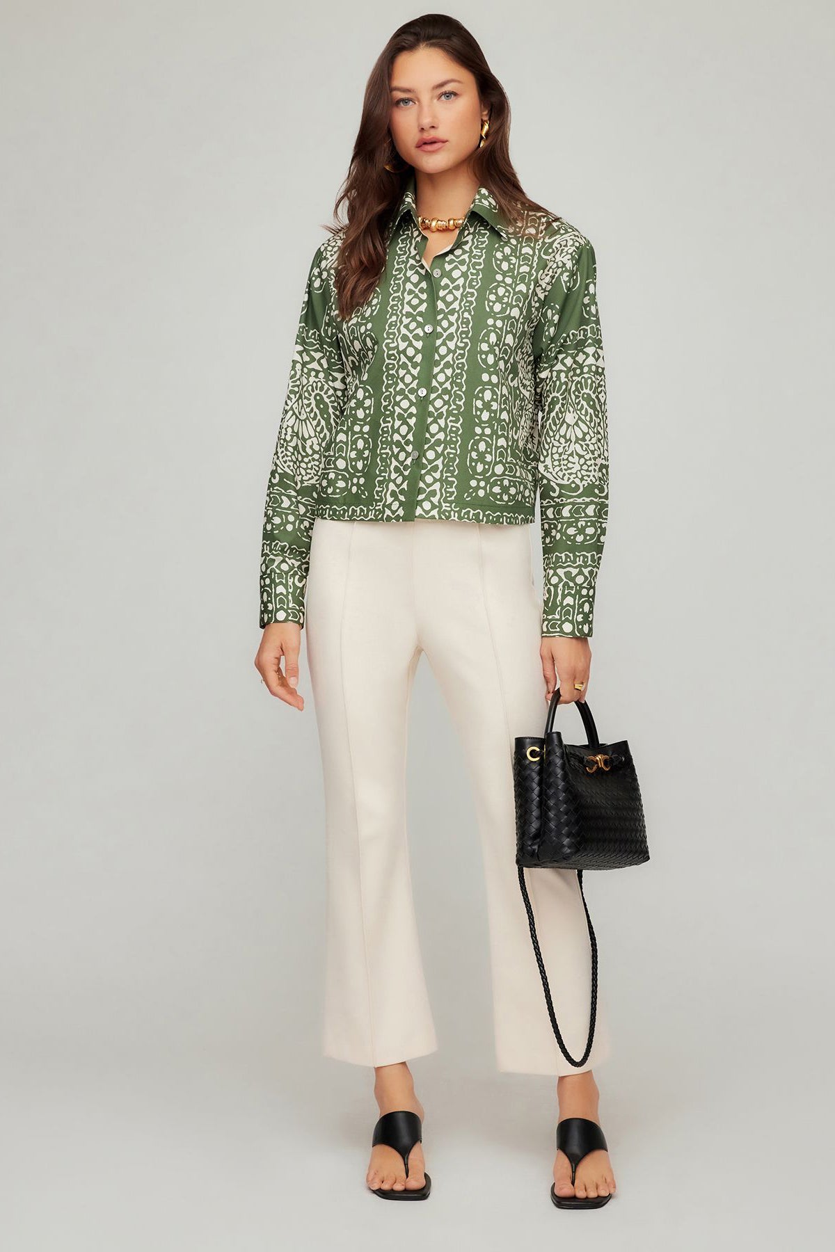 Woman wearing a green patterned blouse and white pants, holding a black handbag on a gray background
