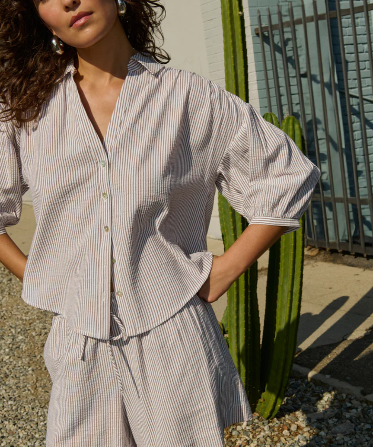 Woman wearing a light gray striped shirt and shorts standing outdoors with a cactus in the background.