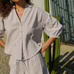 Woman wearing a light gray striped shirt and shorts standing outdoors with a cactus in the background.