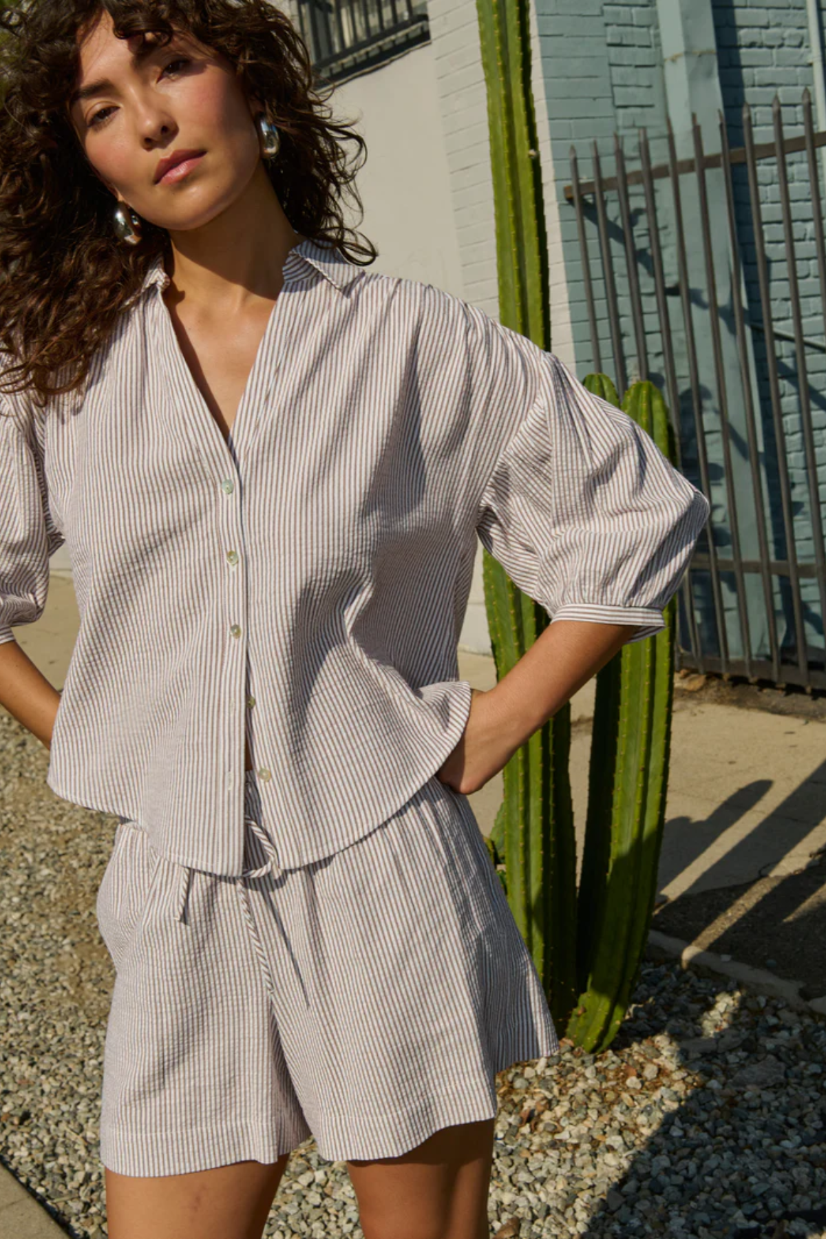 Woman wearing a light gray striped shirt and shorts standing outdoors with a cactus in the background.
