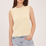 A woman wearing a pale yellow sleeveless knit top and light blue jeans, standing against a plain light beige studio background.