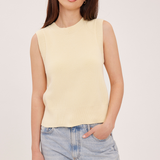 A woman wearing a pale yellow sleeveless knit top and light blue jeans, standing against a plain light beige studio background.