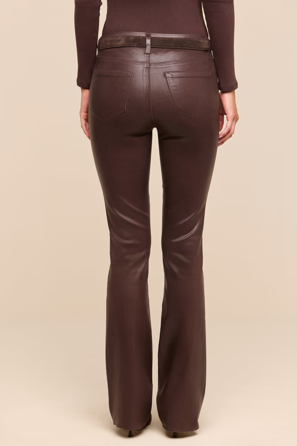 Rear view of a model wearing dark brown faux-leather flare pants with a matching belt, paired with a dark brown ribbed long-sleeve top, against a solid beige background.