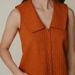 A woman wearing a sleeveless rust-orange ribbed knit top with a wide collar and front buttons, paired with light blue jeans, against a plain beige background.