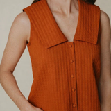A woman wearing a sleeveless rust-orange ribbed knit top with a wide collar and front buttons, paired with light blue jeans, against a plain beige background.