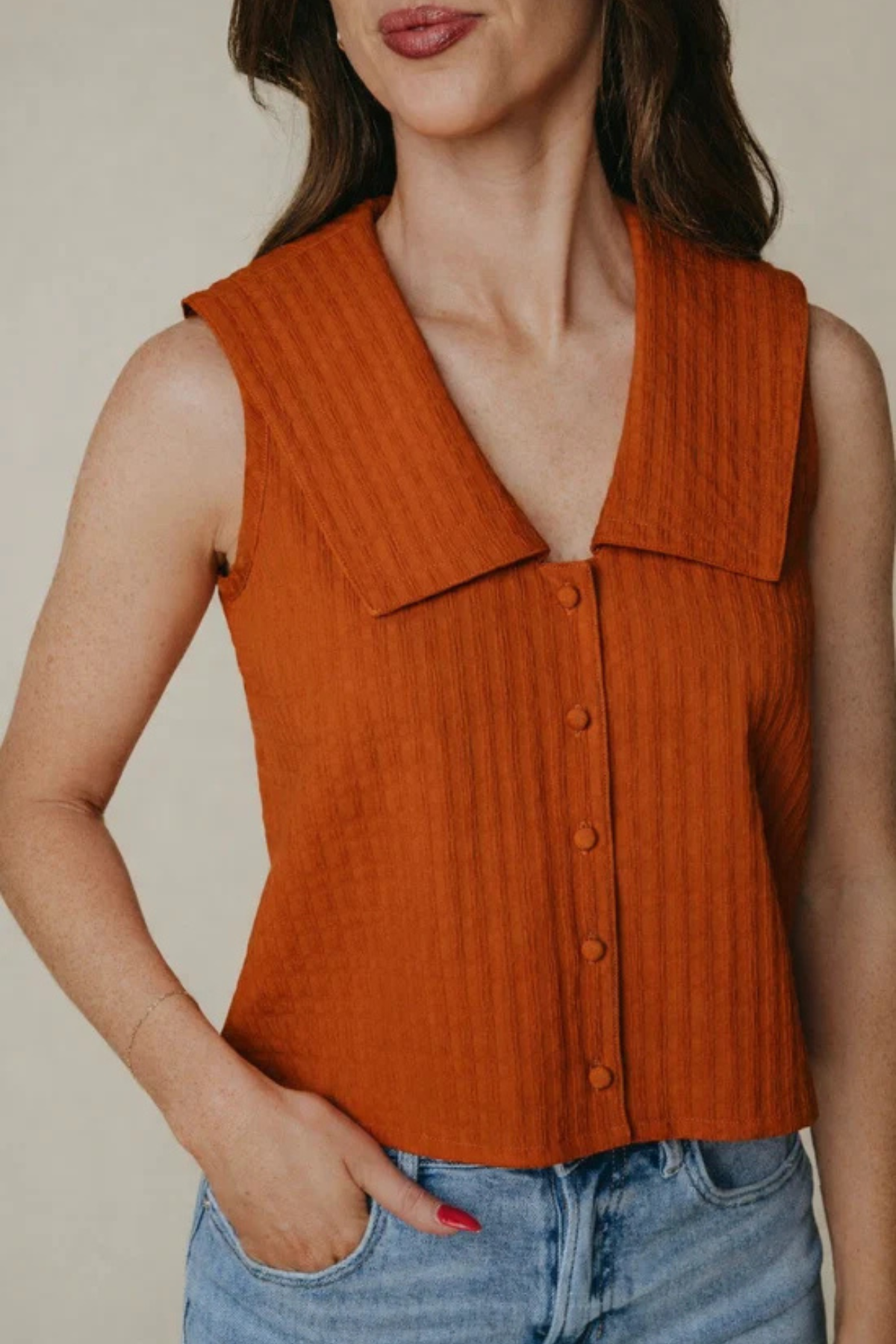 A woman wearing a sleeveless rust-orange ribbed knit top with a wide collar and front buttons, paired with light blue jeans, against a plain beige background.