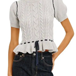 A woman wearing a light gray cable-knit short-sleeve top with puff sleeves, a ruffled hem, and black decorative trim, paired with dark blue jeans against a white background.