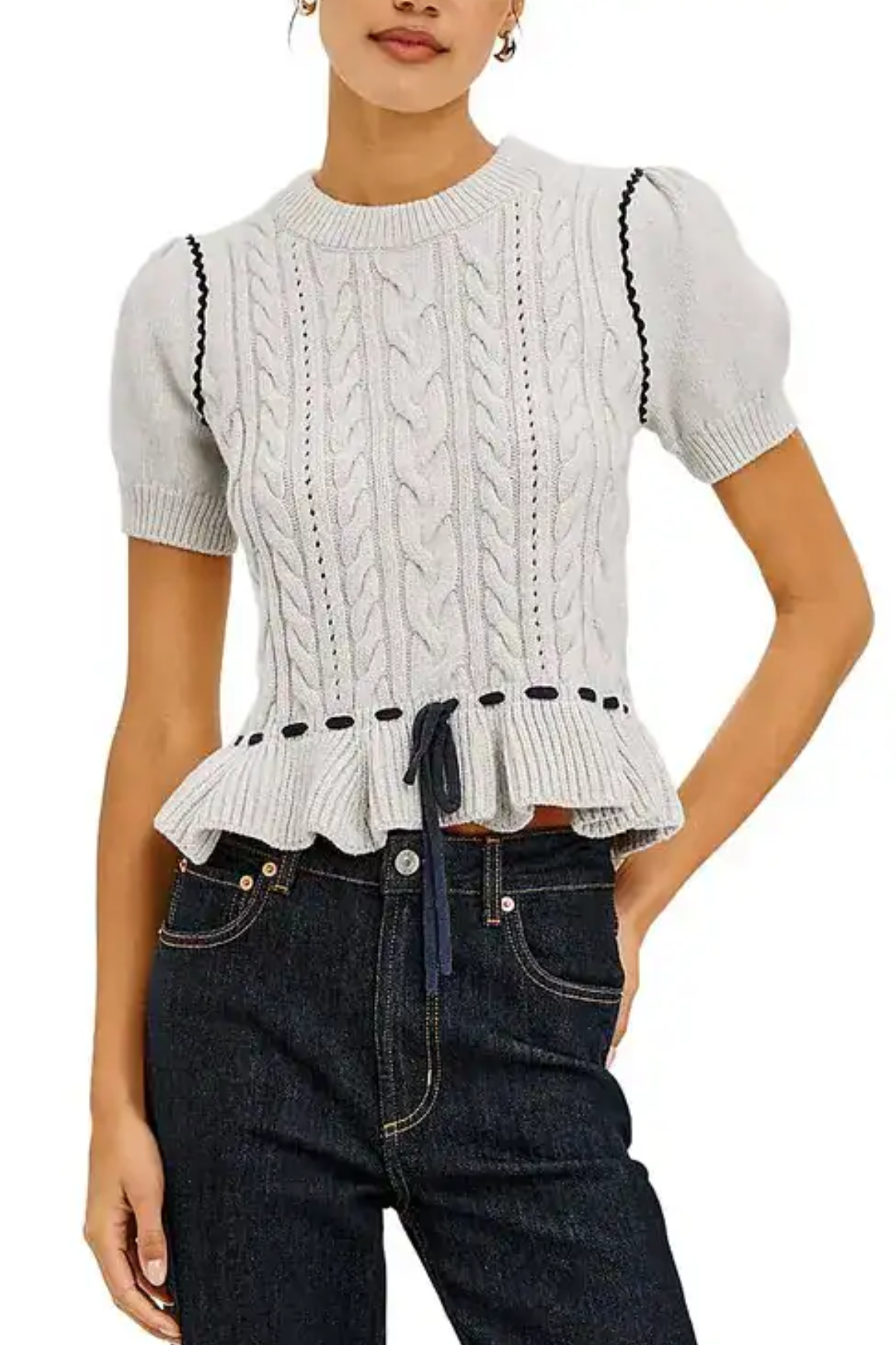 A woman wearing a light gray cable-knit short-sleeve top with puff sleeves, a ruffled hem, and black decorative trim, paired with dark blue jeans against a white background.