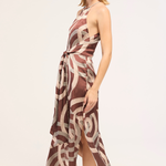 A woman modeling a sleeveless halter-neck midi dress with a brown and beige abstract swirl pattern, shown from a side profile against a plain light background.