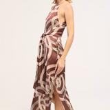 A woman modeling a sleeveless halter-neck midi dress with a brown and beige abstract swirl pattern, shown from a side profile against a plain light background.