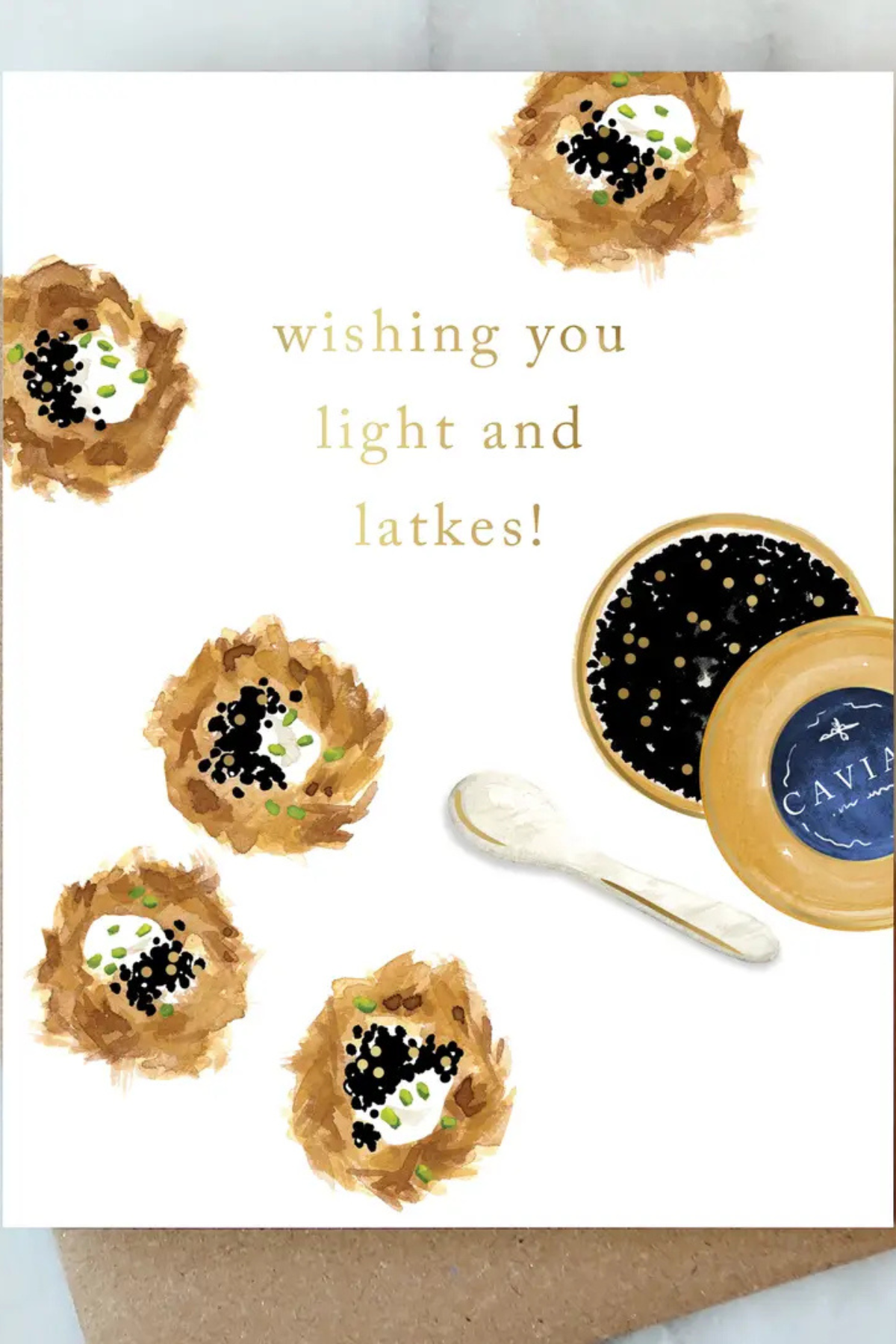 A white greeting card featuring watercolor-style caviar nests and the text 'wishing you light and latkes!' in gold. A small spoon and an open round tin of caviar are pictured beside the text, with a kraft envelope partially visible underneath.
