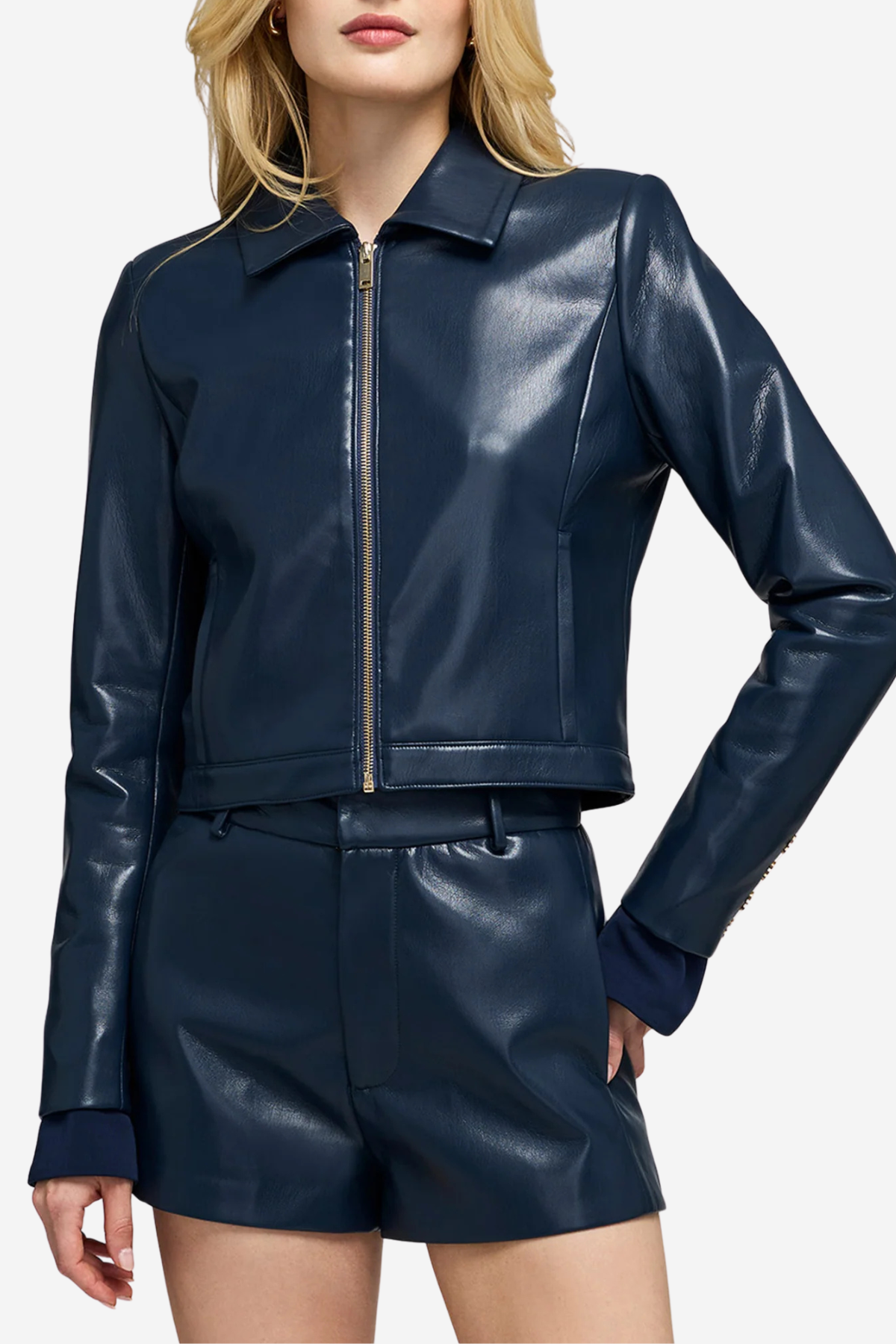 A woman wearing a shiny navy faux-leather cropped jacket with a front zipper and matching navy shorts, shown against a light gray studio background.
