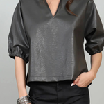 A woman wearing a dark gray faux-leather blouse with puff sleeves and a split V-neck, paired with black pants, against a light gray background.