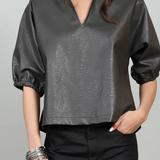 A woman wearing a dark gray faux-leather blouse with puff sleeves and a split V-neck, paired with black pants, against a light gray background.
