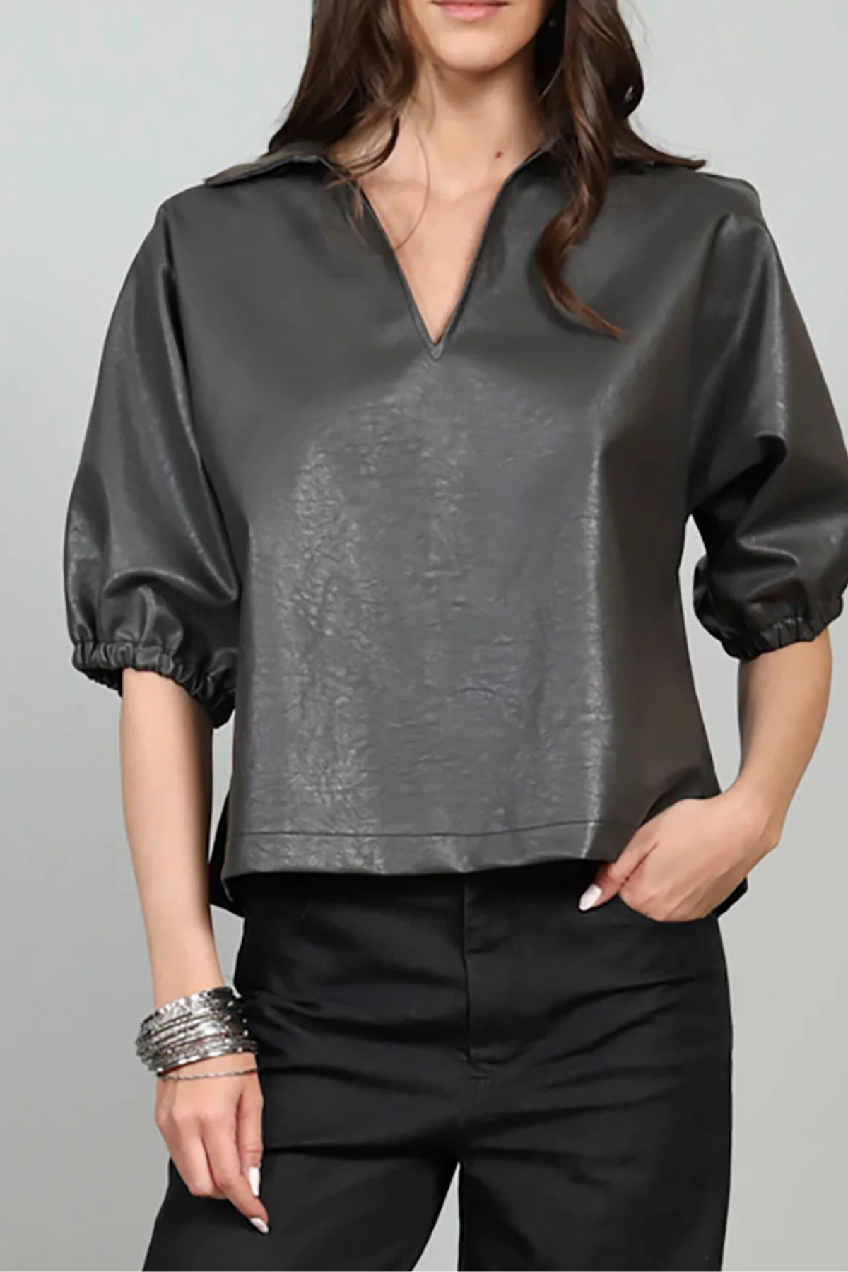 A woman wearing a dark gray faux-leather blouse with puff sleeves and a split V-neck, paired with black pants, against a light gray background.