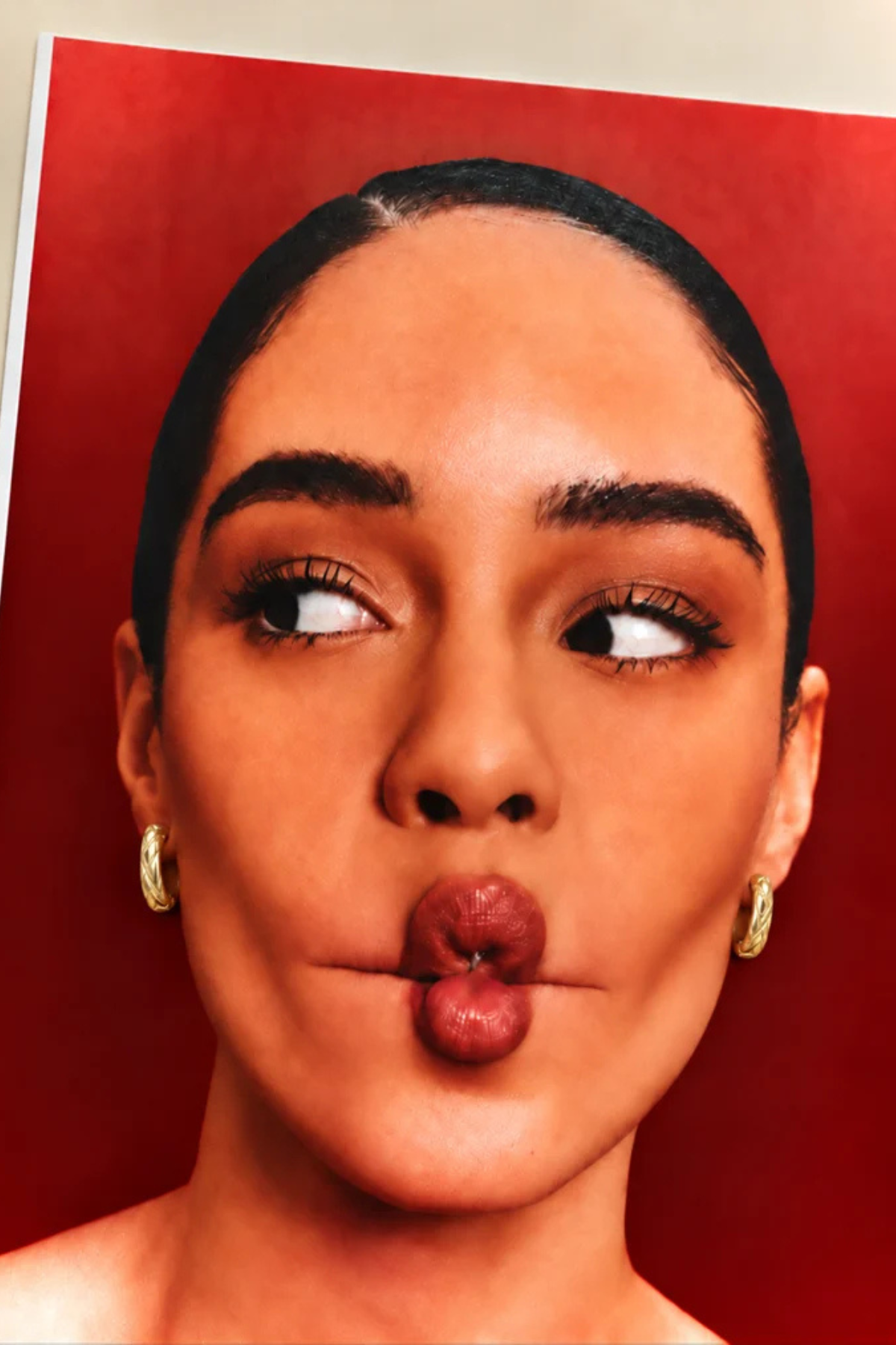 A close-up portrait of a woman with dark hair pulled back, wearing small gold hoop earrings, against a solid red background.