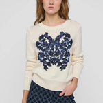 A woman modeling a cream-colored crewneck sweater with a large navy blue floral embroidery design on the front, paired with dark patterned trousers, against a light gray studio background.