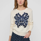 A woman modeling a cream-colored crewneck sweater with a large navy blue floral embroidery design on the front, paired with dark patterned trousers, against a light gray studio background.