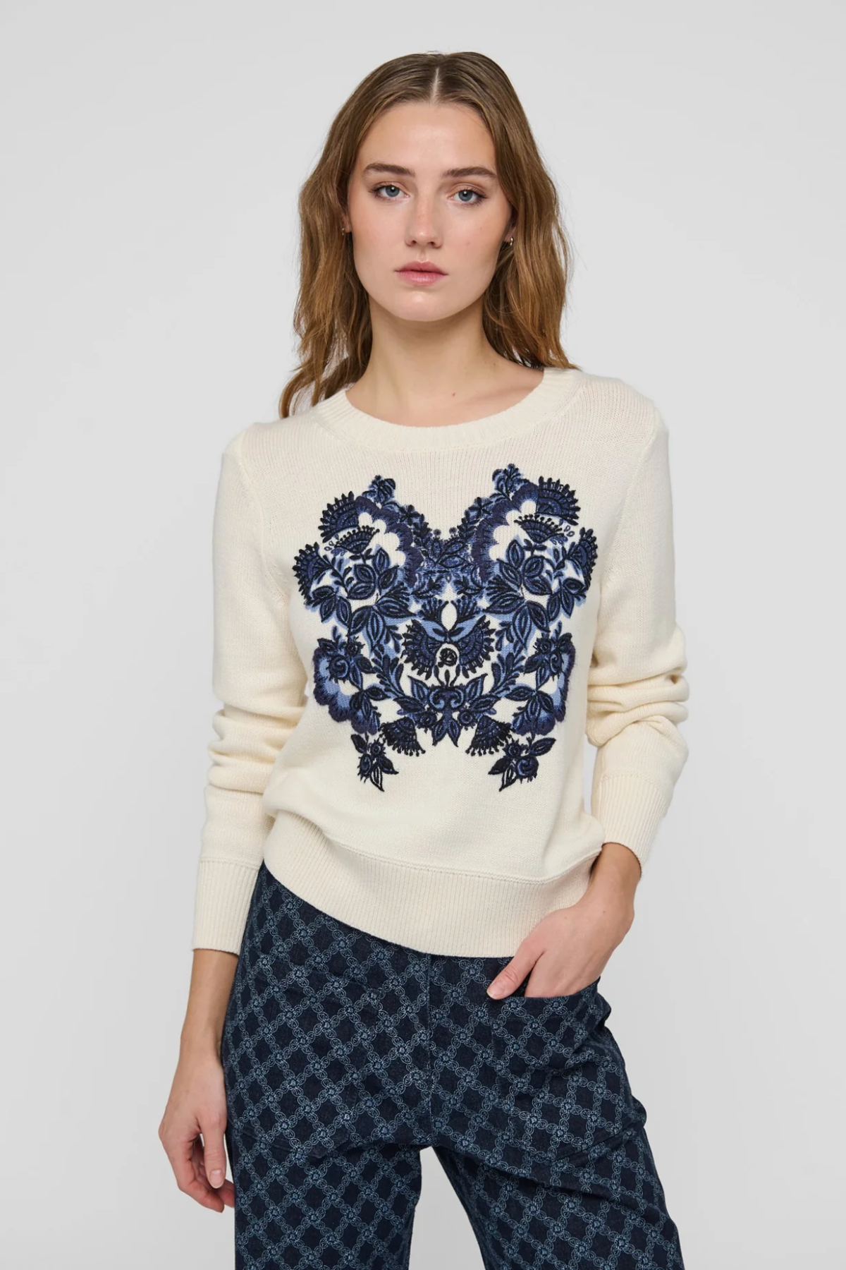 A woman modeling a cream-colored crewneck sweater with a large navy blue floral embroidery design on the front, paired with dark patterned trousers, against a light gray studio background.