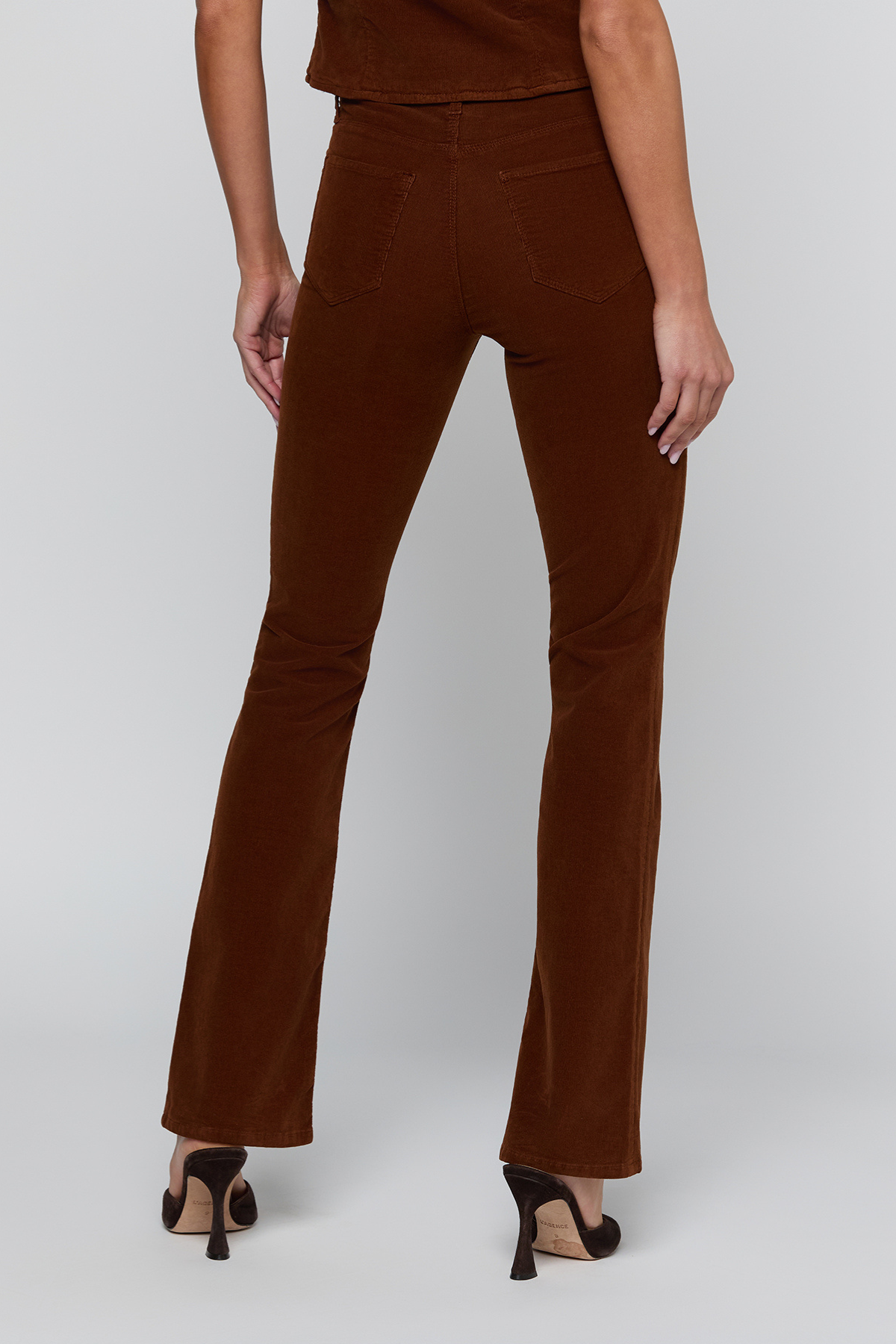 Rear view of a model wearing brown corduroy flare pants and black high heels against a light gray studio background.