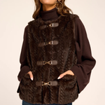 A woman wearing a dark brown faux fur vest with gold-tone buckle closures over a dark brown turtleneck sweater and blue jeans, photographed against a plain light beige studio background.