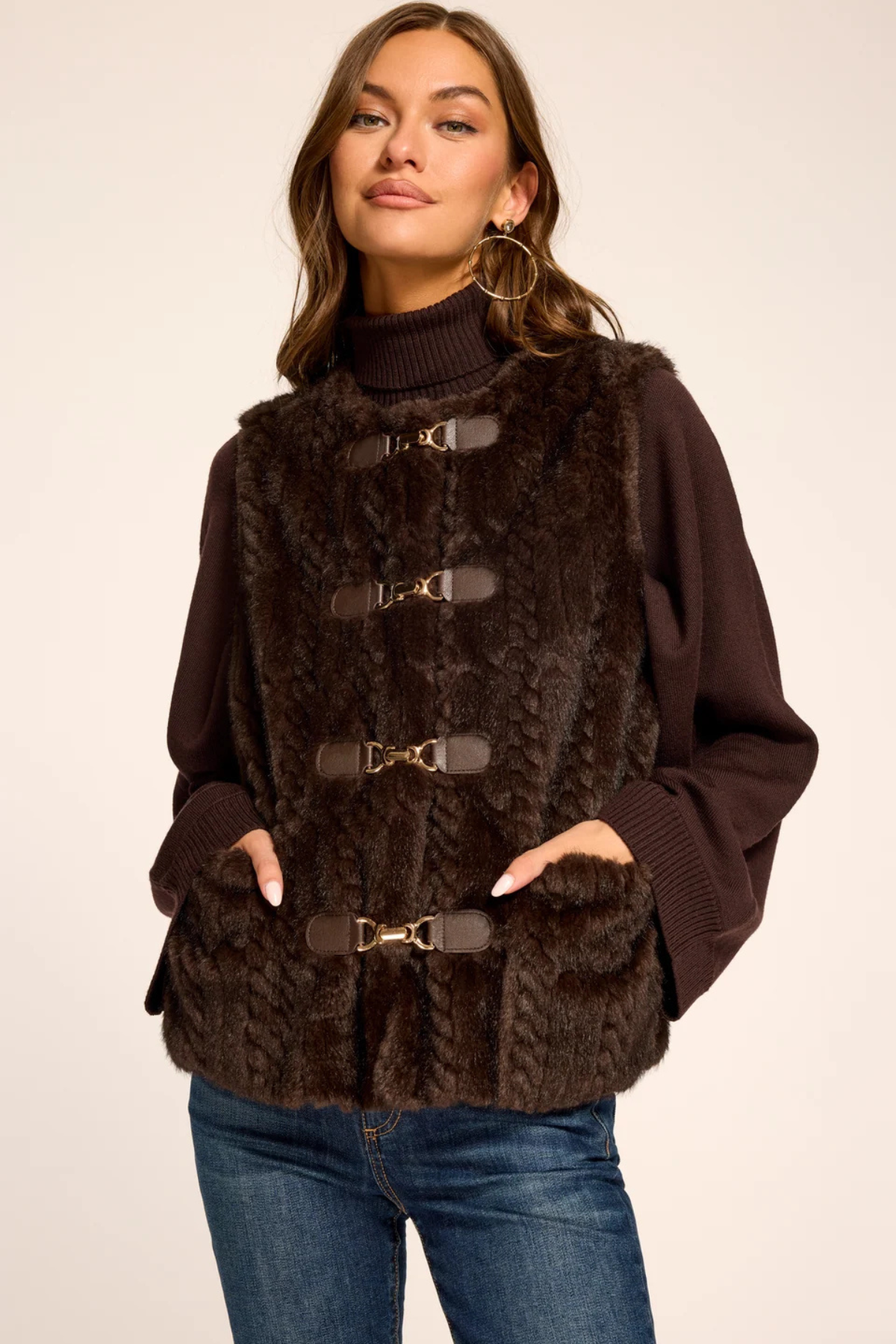 A woman wearing a dark brown faux fur vest with gold-tone buckle closures over a dark brown turtleneck sweater and blue jeans, photographed against a plain light beige studio background.