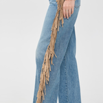 A side-profile view of a person wearing light blue wide-leg jeans with long tan fringe hanging down the outer side, paired with snakeskin-patterned heels against a plain light gray studio background.