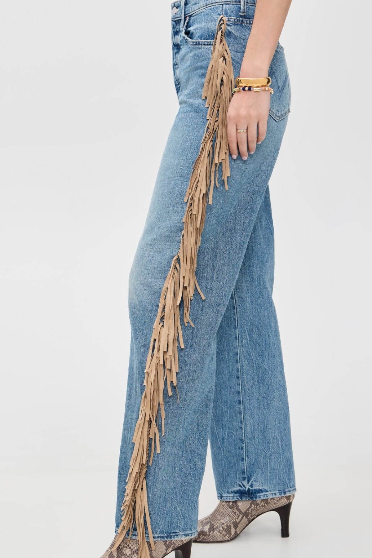 A side-profile view of a person wearing light blue wide-leg jeans with long tan fringe hanging down the outer side, paired with snakeskin-patterned heels against a plain light gray studio background.